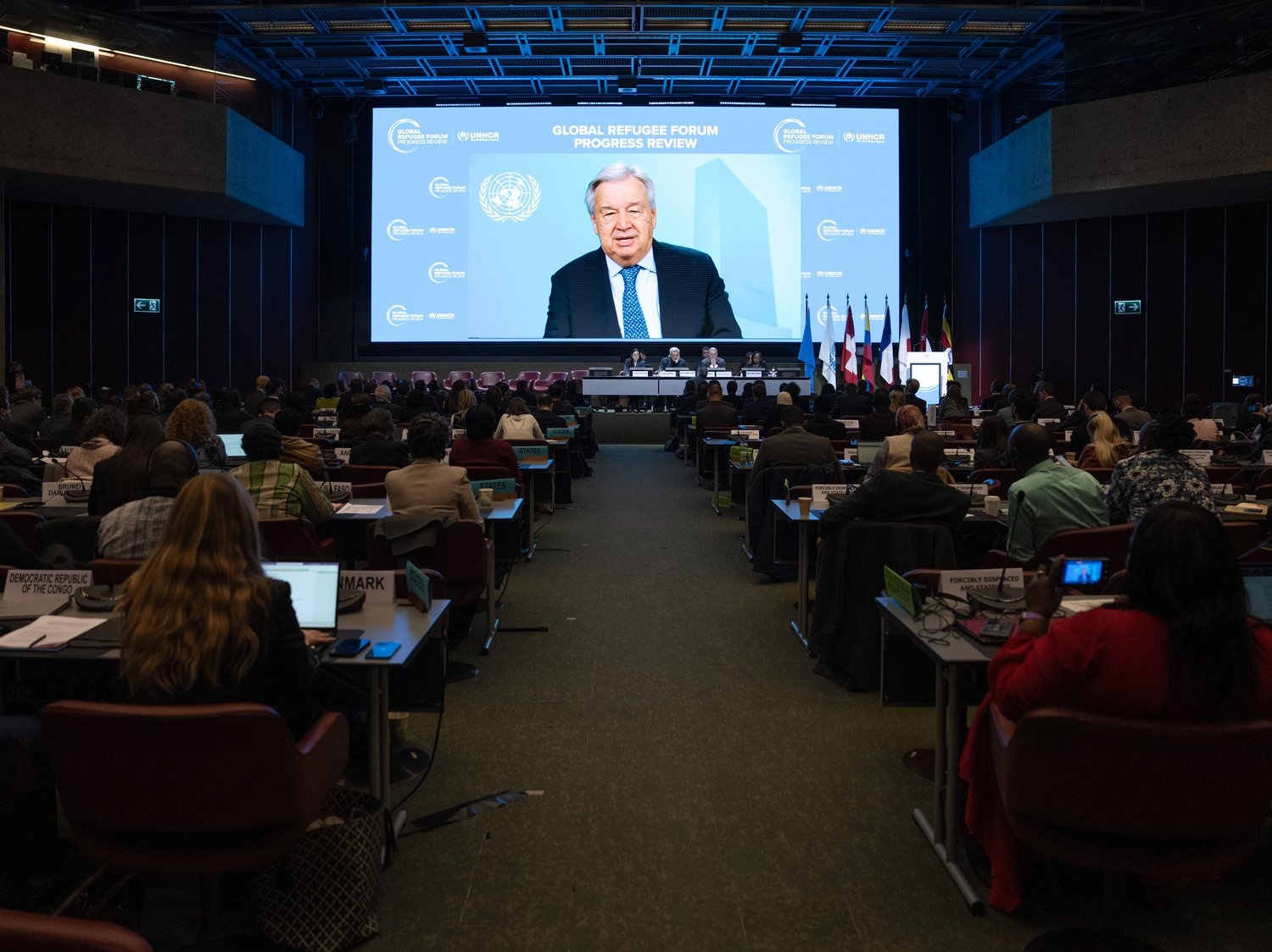 A man in a suit is shown on a giant screen in a darkened conference room