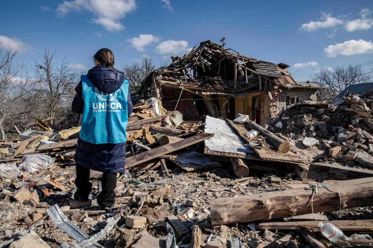 A woman wearing a blue UNHCR vest stands in front of the remains of a destroyed house