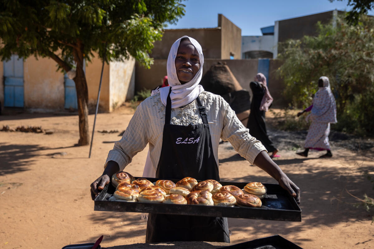 A woman wearing an apron holds a tray of bread and pastries.