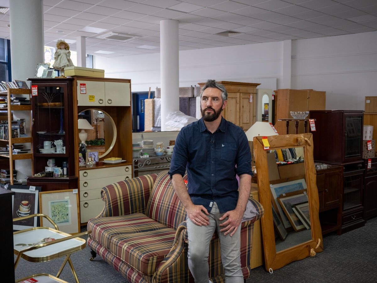 A man wearing a blue shirt and jeans leans on the arm of a sofa in a second-hand furniture store.