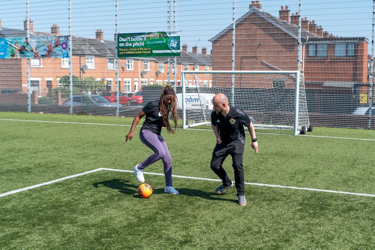 A woman and a man play football on a pitch with houses in the background