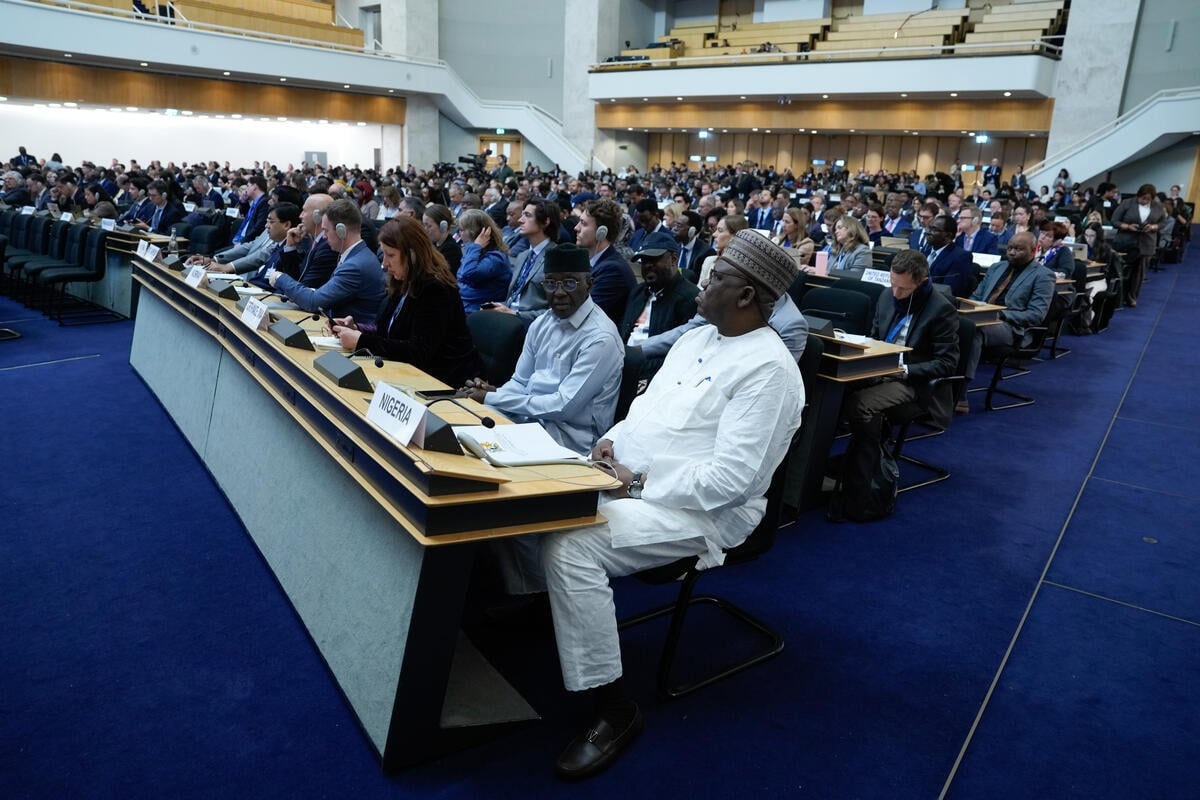 Rows of people in a large auditorium are seated behind long desks with country names displayed in front of them 