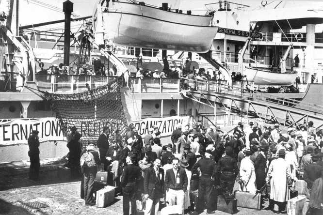 Black and white photo: A group fo refugees wait to board a ship 