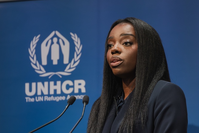 A photo of Jenny BSG speaking into microphones next to the UNHCR logo