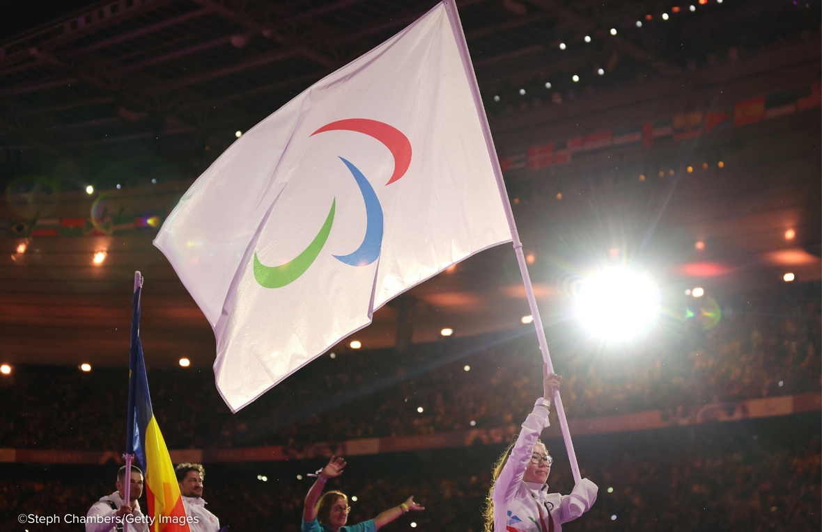 A woman wearing glasses holds a large IPC flag above her head in a brightly lit stadium.