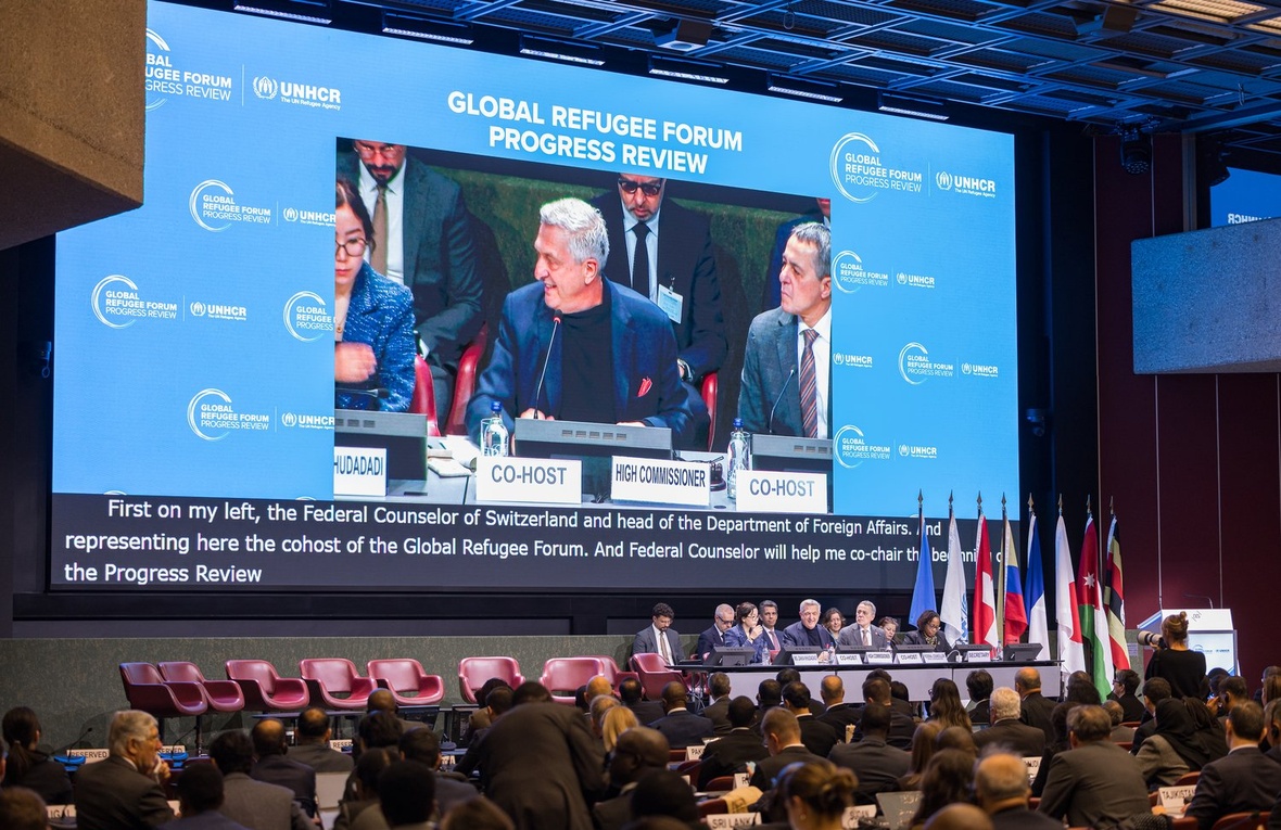 A man is pictured speaking on a giant screen in front of a conference room audience