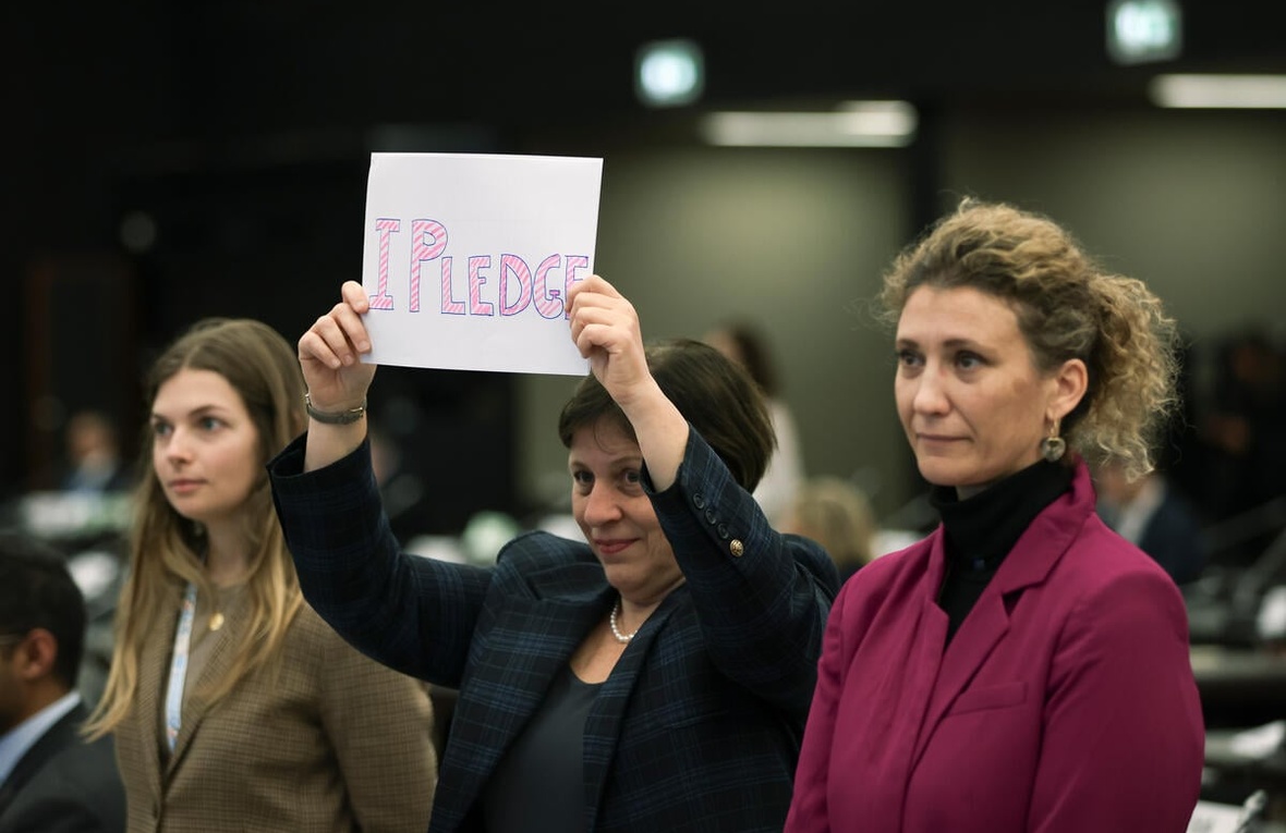 Three individuals stand together, one holds up a sign that reads 'I pledge'.
