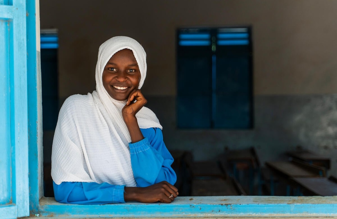 A young girl smiles at the camera, looking through an open window.