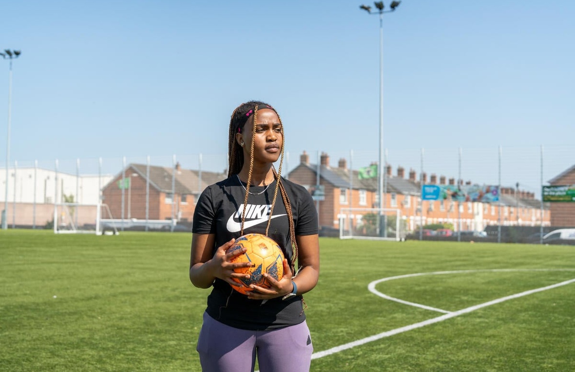 A young woman holds a football on a football pitch with rows of houses in the background