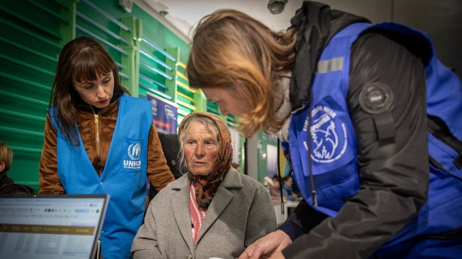 Two aid workers stand beside an elderly woman seated in front of a desk