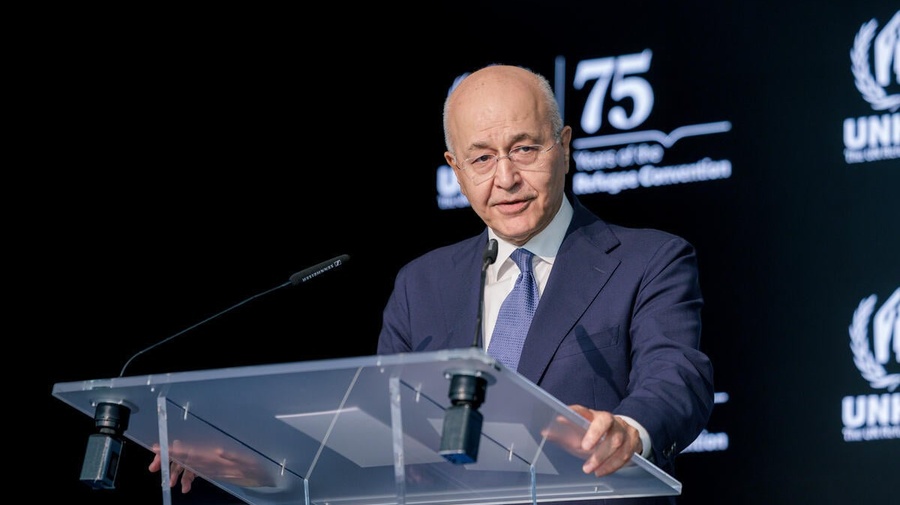 A man in a suit delivers a speech standing at a clear lectern in front of a dark background.