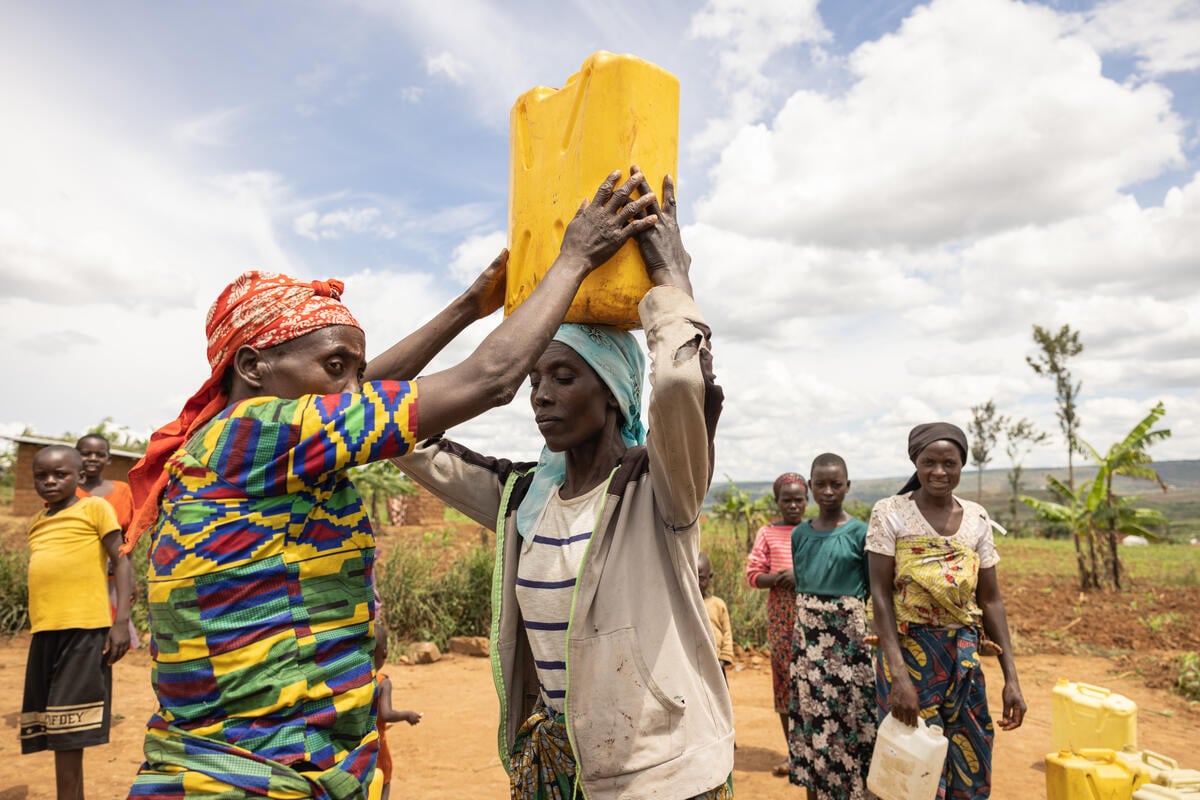 Dans un paysage aride, des femmes aux vêtements colorés collectent de l'eau dans des bidons qu'elles portent sur leur tête ou dans leurs mains.