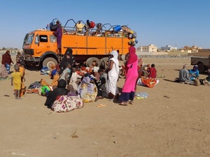 Des familles déracinées se rassemblent avec leurs affaires devant et sur un grand camion, dans un environnement aride.
