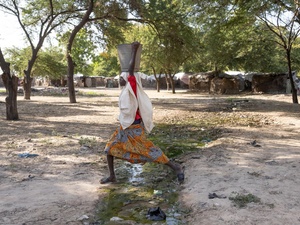 Une jeune fille portant un seau sur la tête enjambe un ruisseau qui traverse un camp.