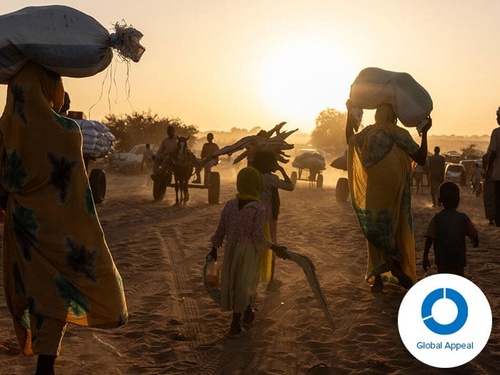 Women and children walk through a dusty field, balancing firewood and white hessian sacks on their heads, silhouetted against the setting sun.