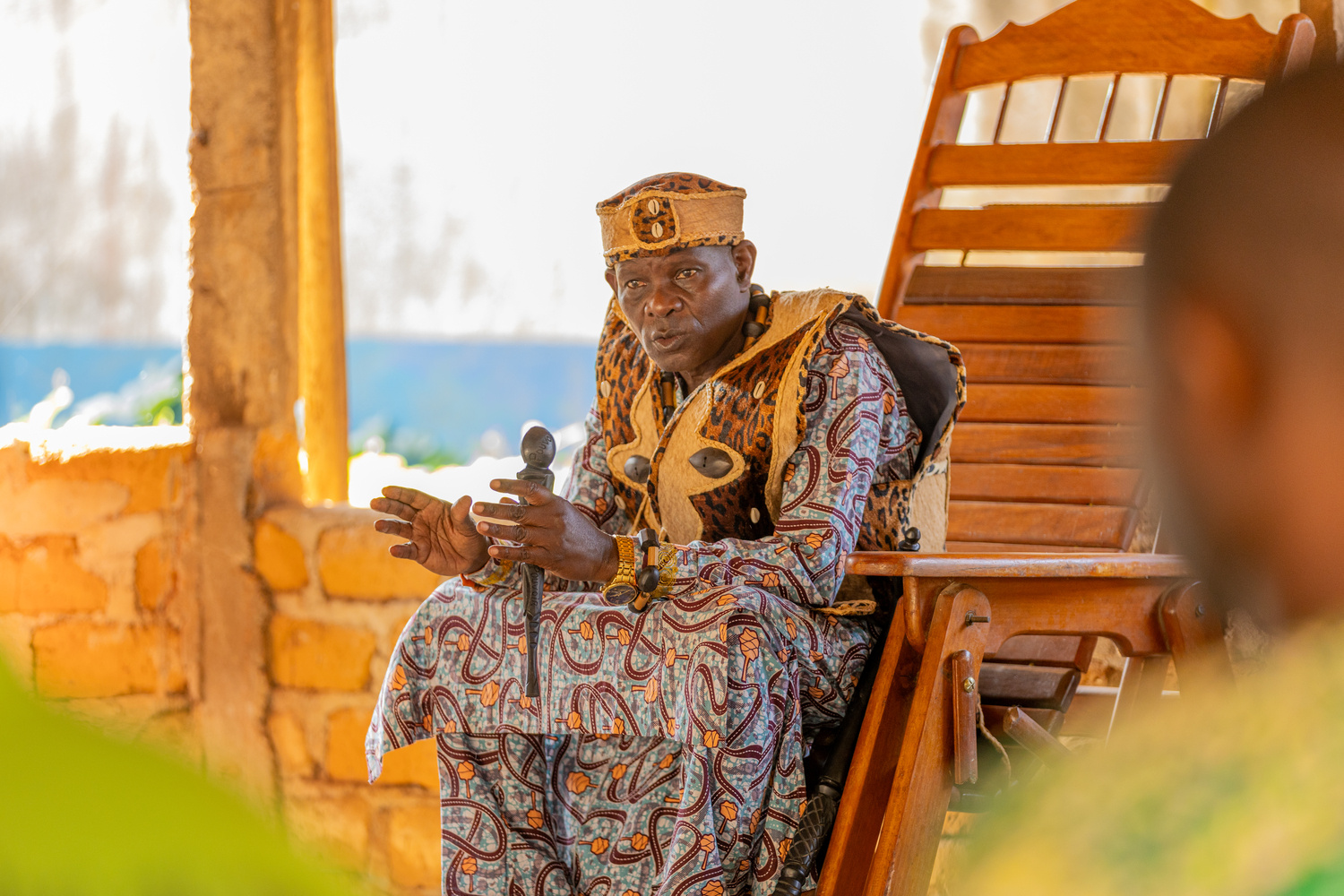 A man in colourful robes sits in a large wooden chair and speaks 
