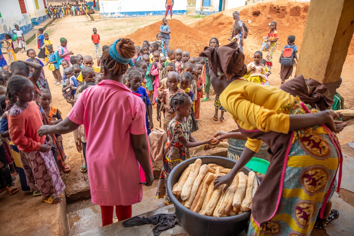 Women wearing aprons distribute bread from a large container to a group of children