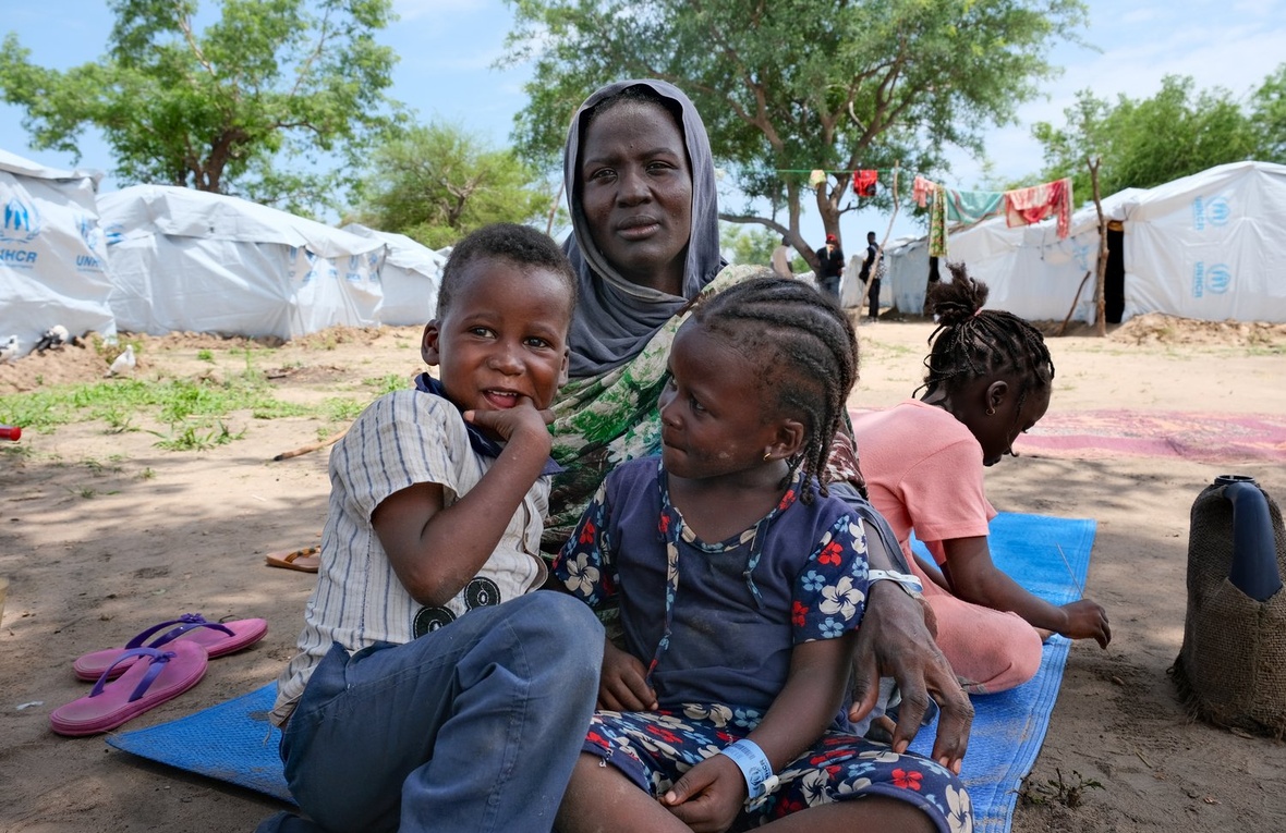 Une femme est assise sur une natte à l'extérieur avec trois jeunes enfants. Des tentes du HCR sont visibles en arrière-plan.