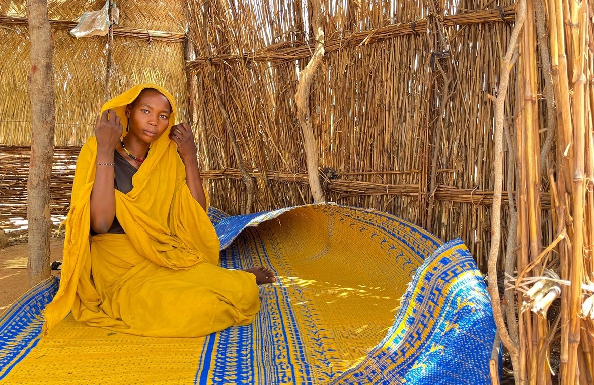 Une femme vêtue de jaune est assise sur une natte et ajuste son foulard dans un abri de paille.