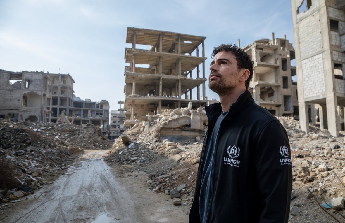 A man wearing a black UNHCR jacket looks upwards while surrounded by rubble and destroyed buidings