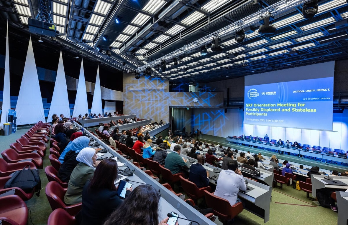 Les participants à l'immense amphithéâtre du Forum mondial sur les réfugiés 2023 regardent une diapositive indiquant "GRF Orientation Meeting for Forcibly Displaced and Stateless Participants" (Réunion d'orientation du GRF pour les participants déplacés de force et apatrides).