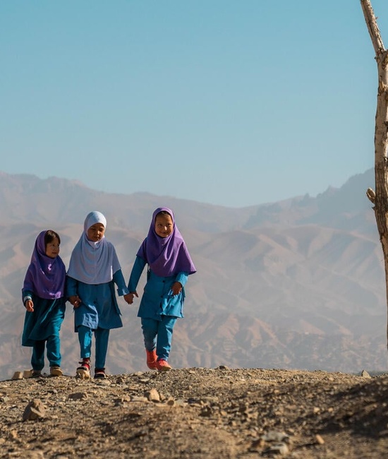 Trois jeunes réfugiées afghanes marchent main dans la main sur un sol aride, une chaîne de montagnes derrière elles.
