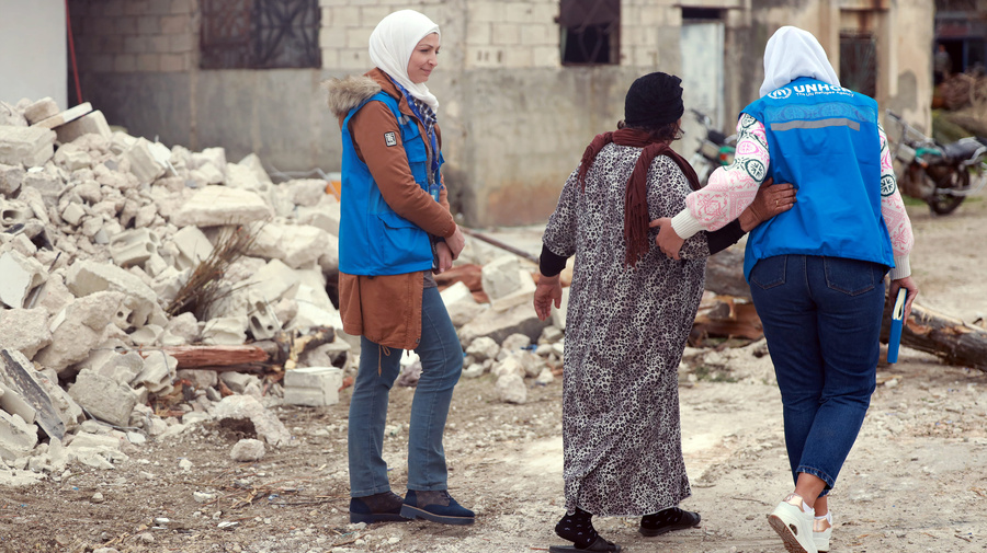 Deux employées du HCR marchent avec une personne déplacée syrienne à côté des débris de sa maison.