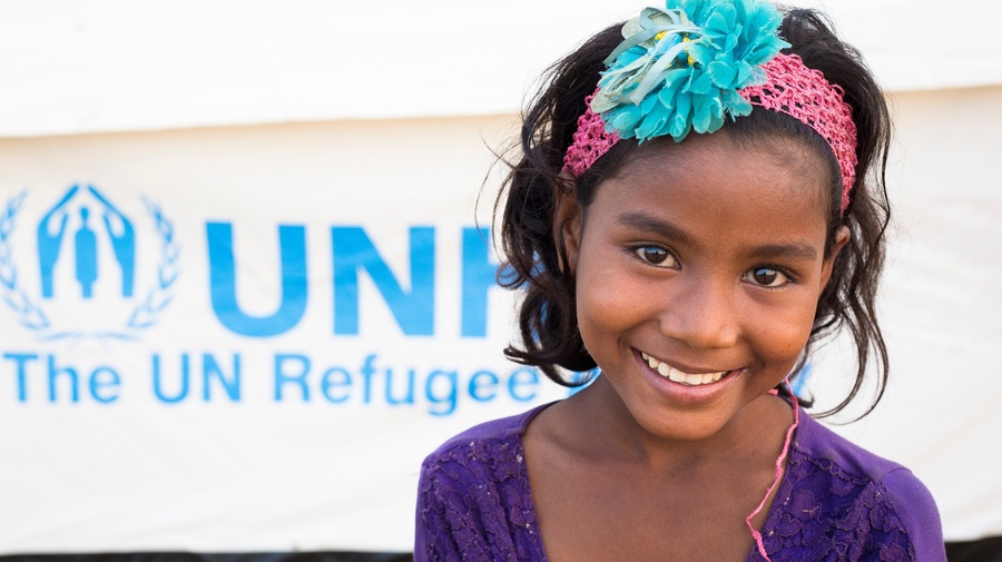 Une petite fille sourit devant une tente au logo du HCR.