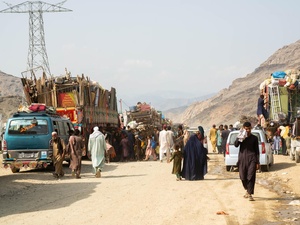 Afghan returnees walk along a dirt road beside loaded trucks.