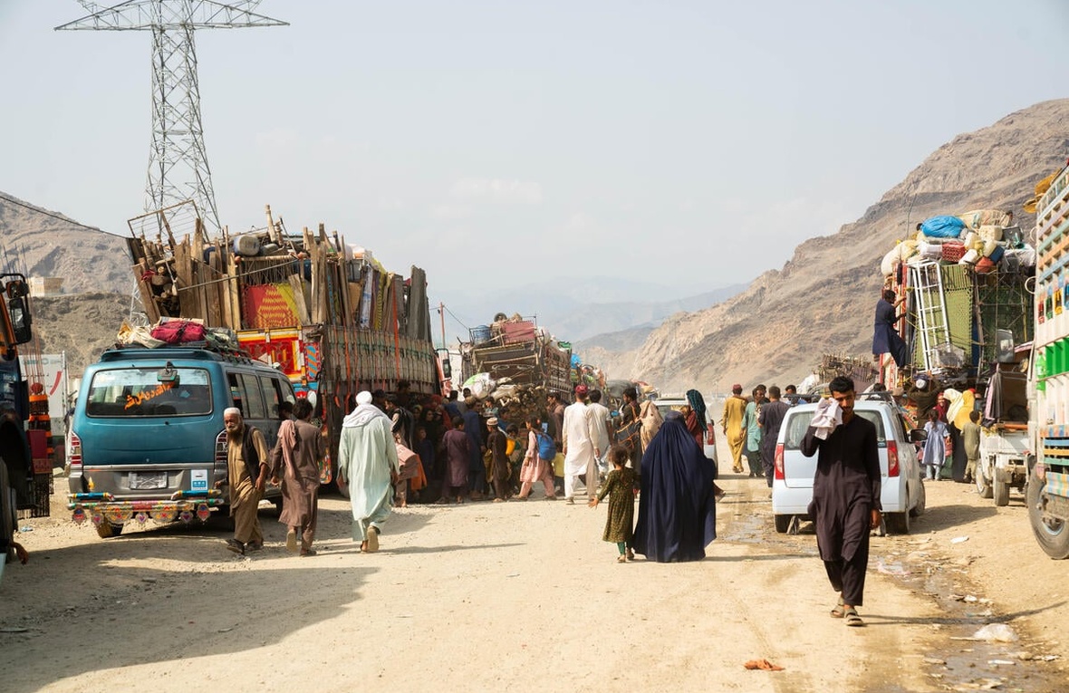 Afghan returnees walk along a dirt road beside loaded trucks.