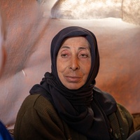 A woman in a dark headscarf sitting inside a tent looks directly at another woman in the foreground wearing a blue jacket and white headscarf whose face is turned away