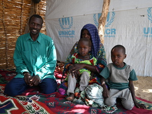 A couple sit on a carpet inside a woven reed shelter accompanied by two young children