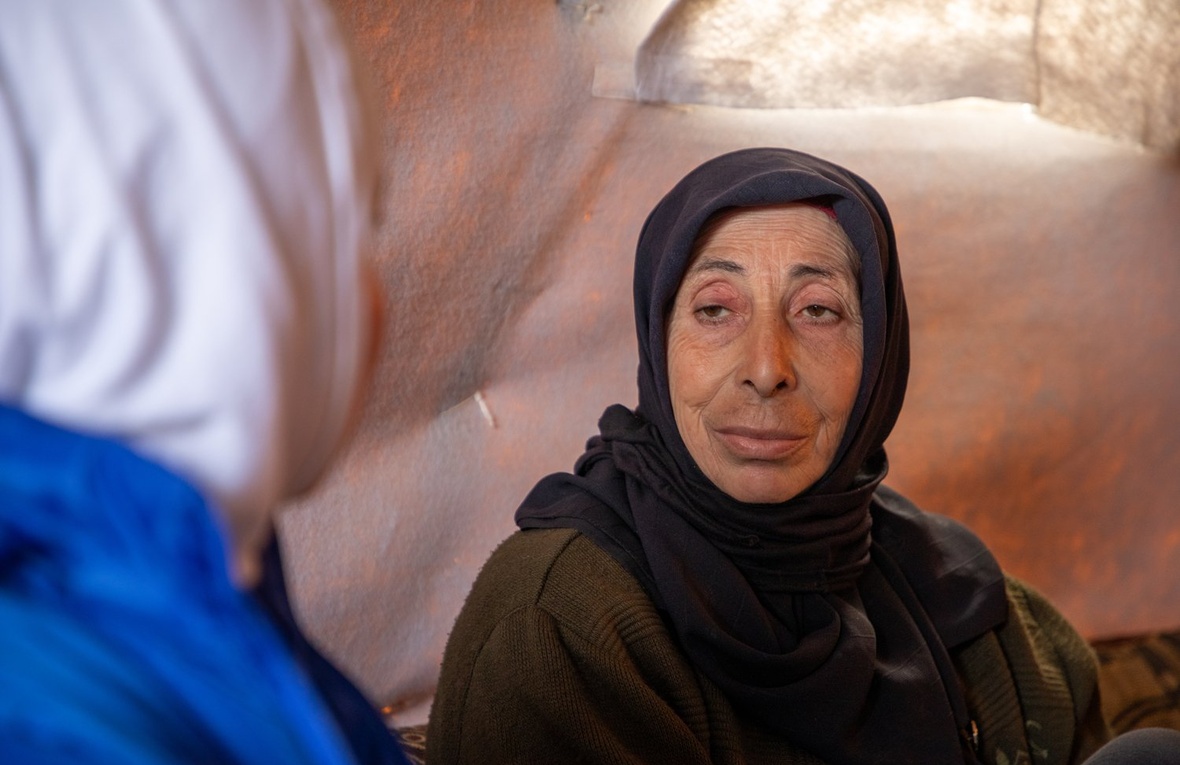 A woman in a dark headscarf sitting inside a tent looks directly at another woman in the foreground wearing a blue jacket and white headscarf whose face is turned away