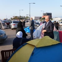 A man in a suit speaks to a family in an open area filled with tents and vehicles 