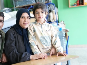 A couple and their daughter sit at a school desk in a classroom with a bed in the background 