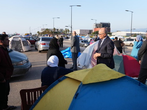 A man in a suit speaks to a family in an open area filled with tents and vehicles 