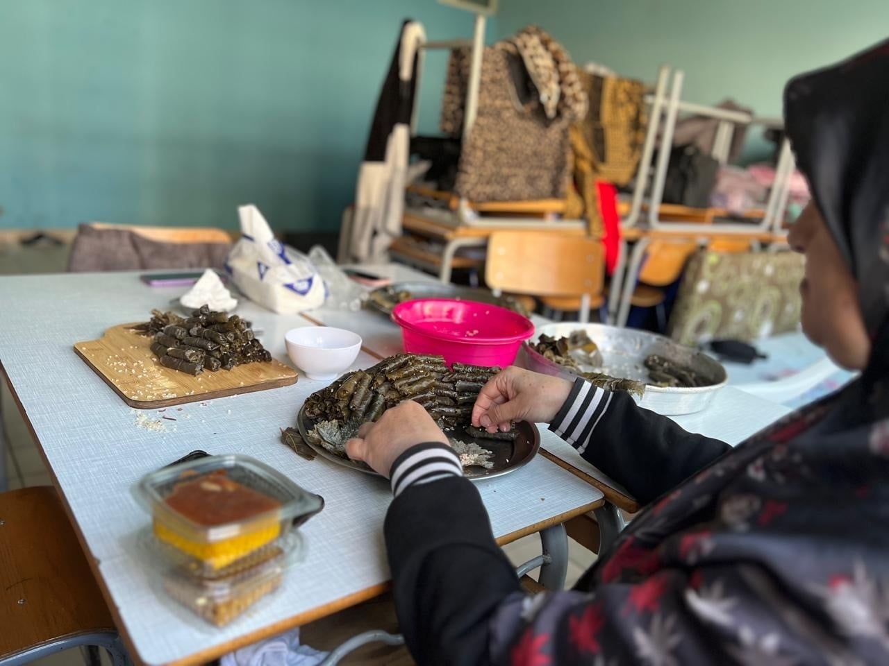 A woman sits at a school desk stuffing vine leaves with rice and arranging them on a plate
