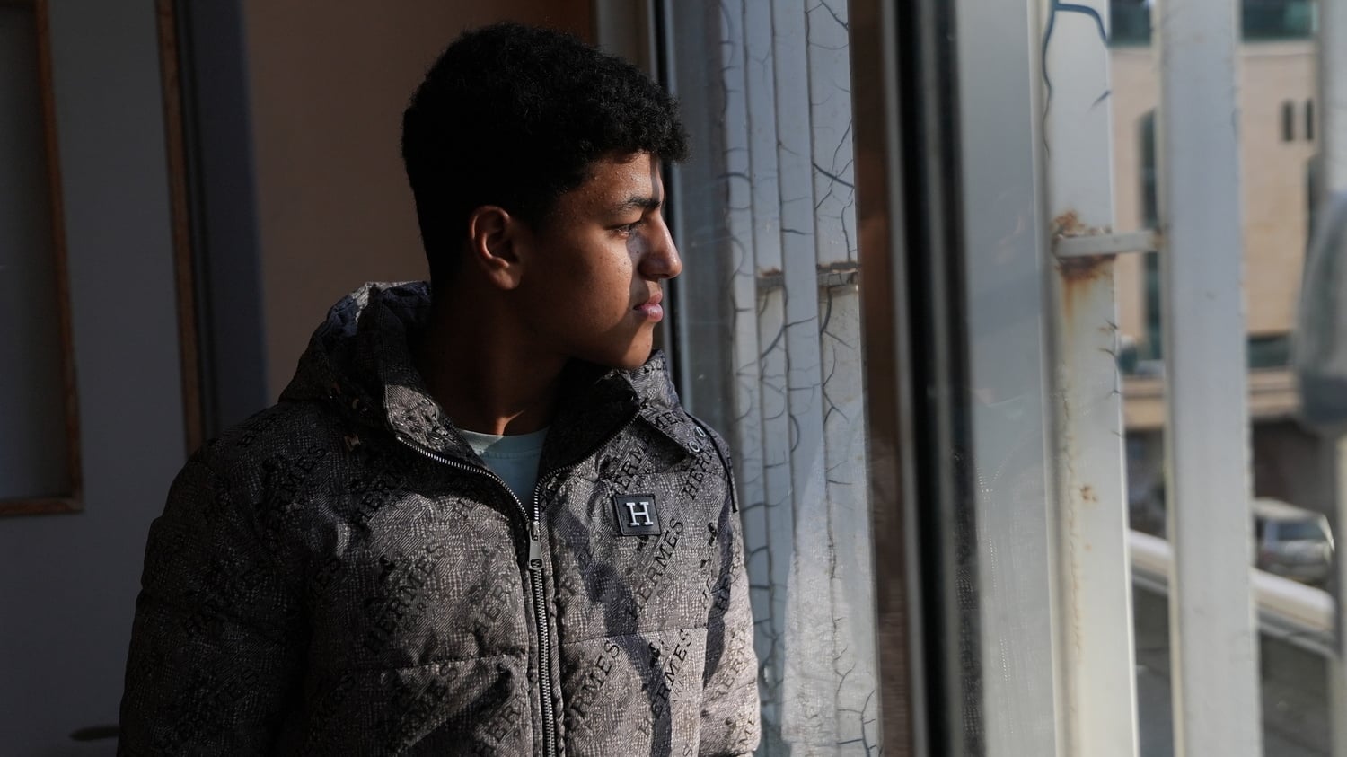 A young man stands next to a window looking out onto a courtyard