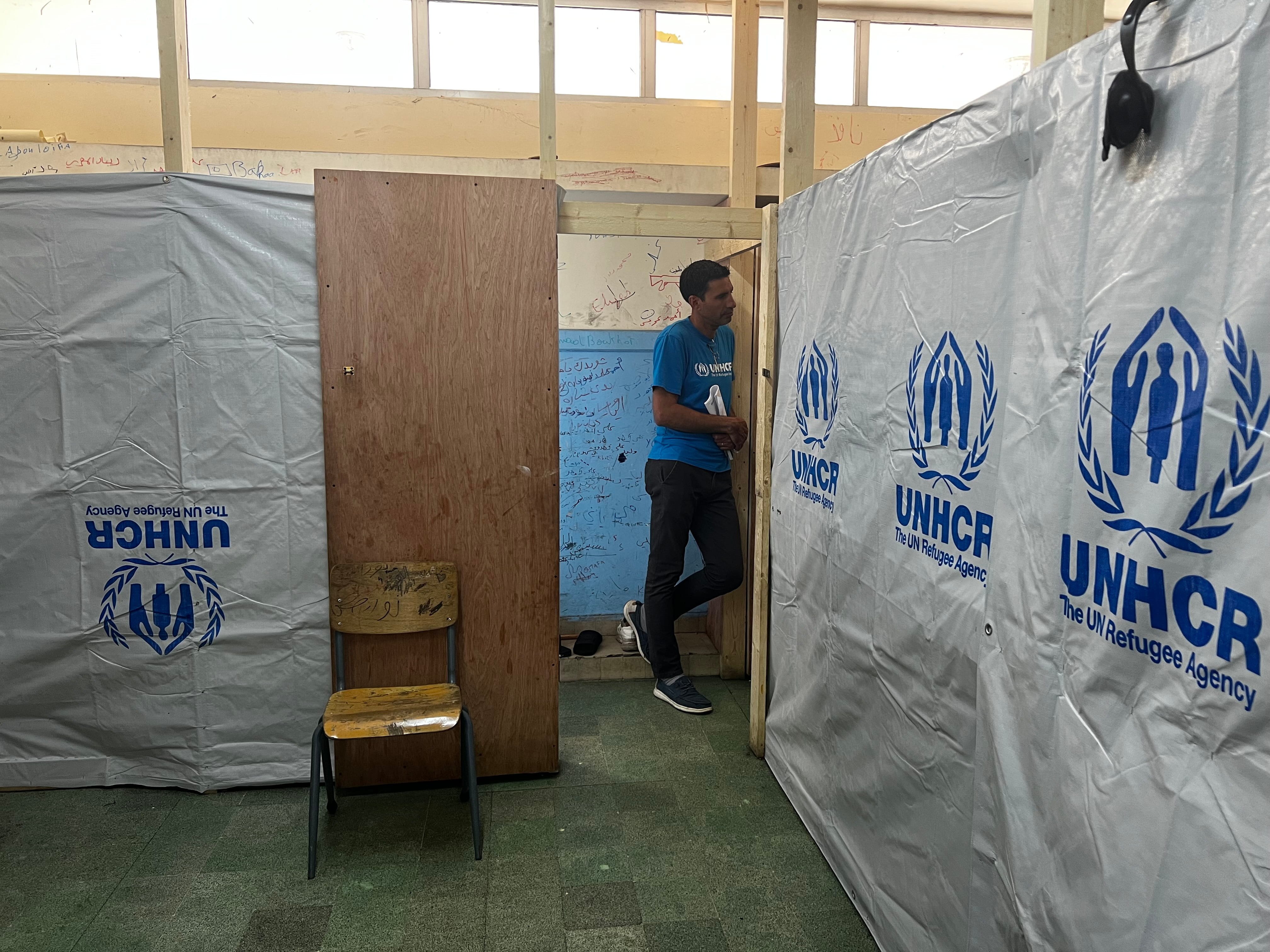 A man wearing a blue t-shirt and black trousers leans in a doorway inside a room partitioned with wooden frames and UNHCR plastic sheeting