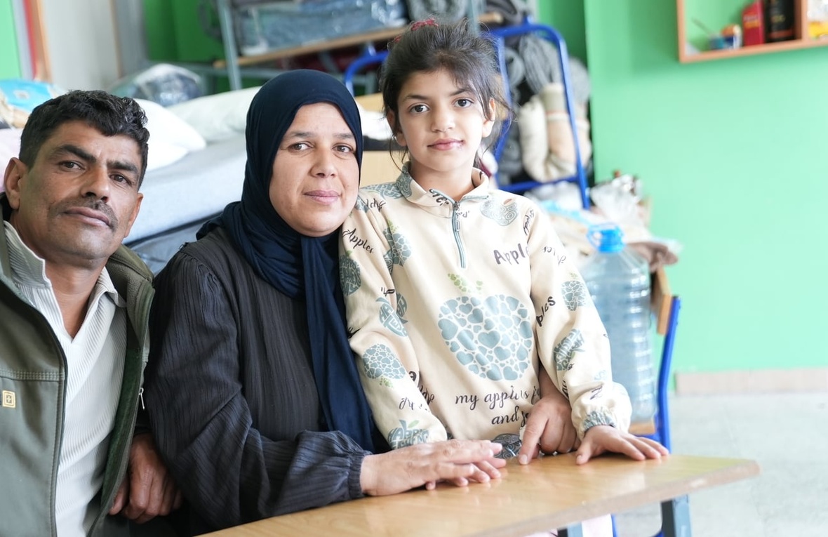 A couple and their daughter sit at a school desk in a classroom with a bed in the background 