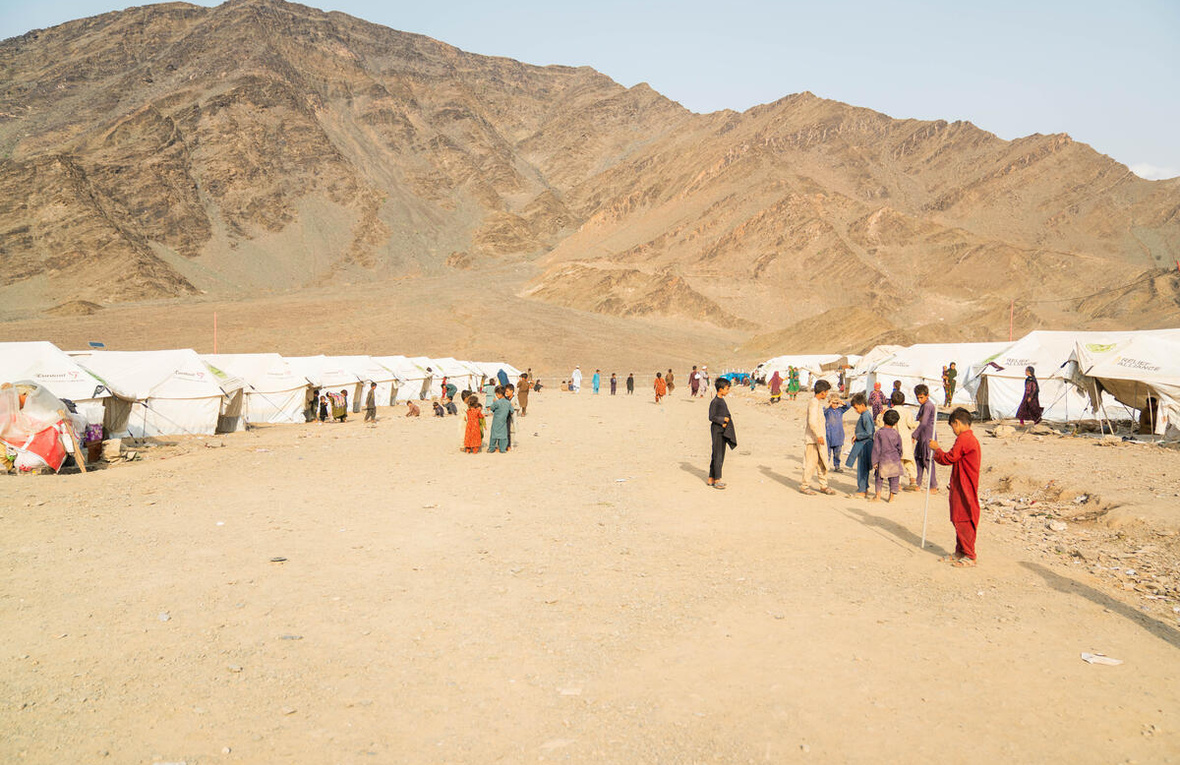 Tents line a dirt road where children gather, with a mountain in the background