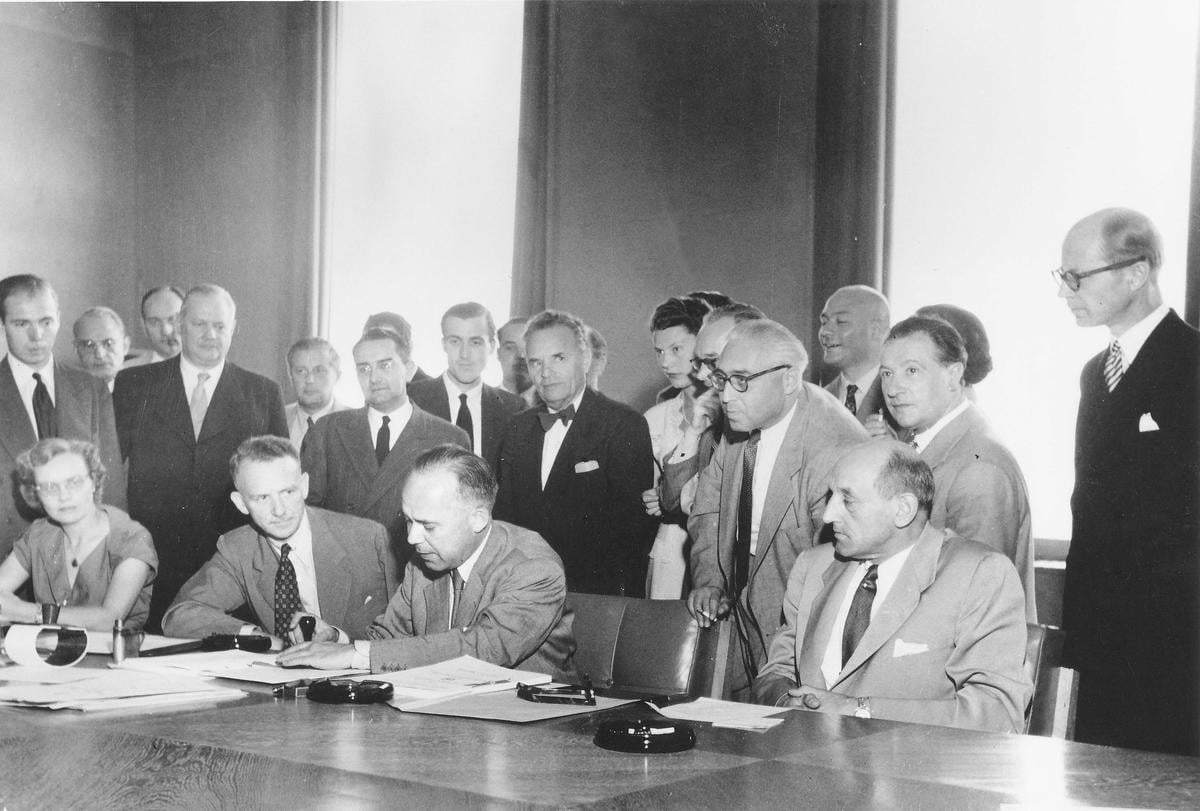 Back and white photo: A man signs the 1951 Refugee Convention, surrounded by a group of people.