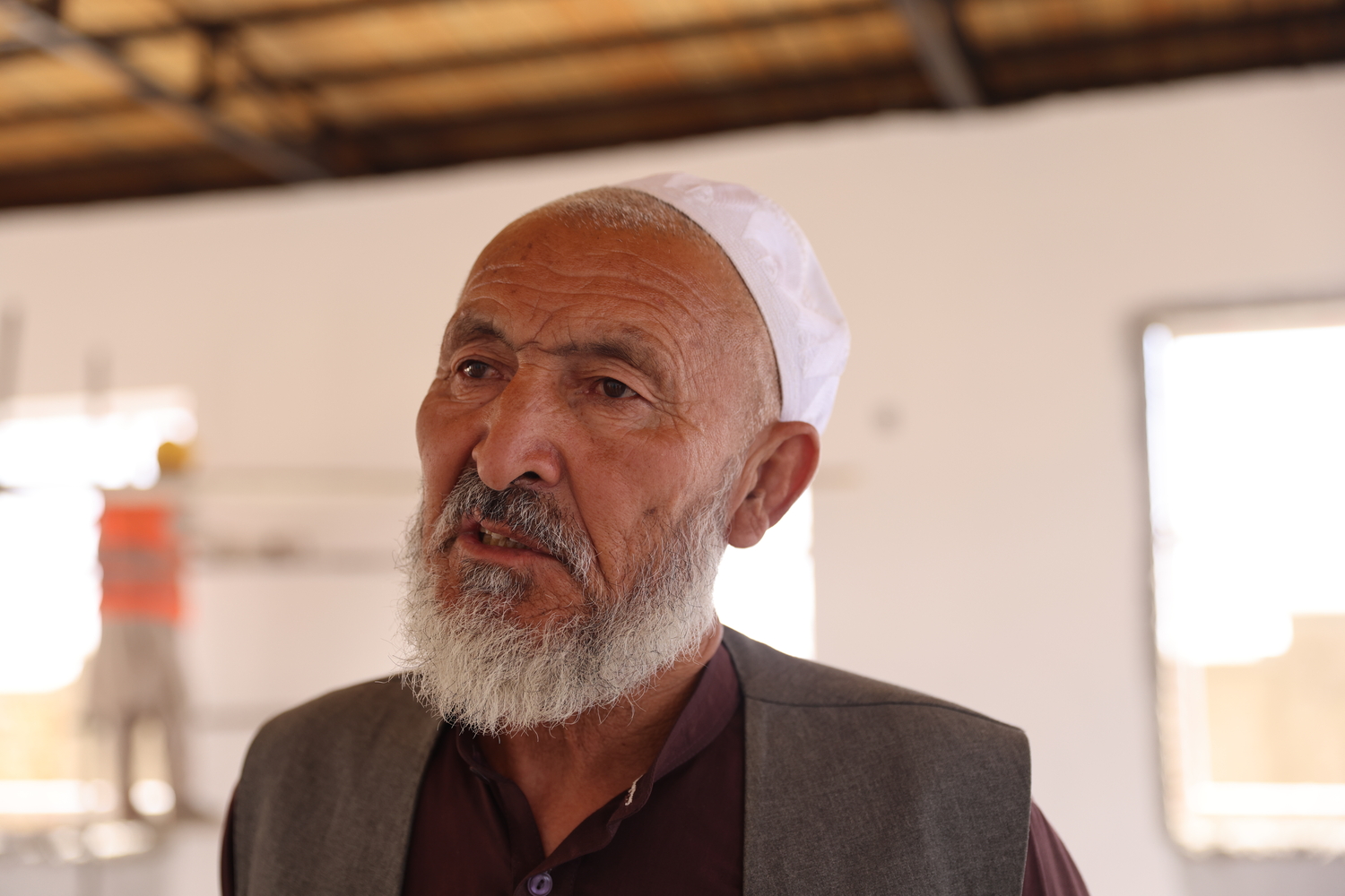 An older man with a beard and white fez speaks to someone off camera.