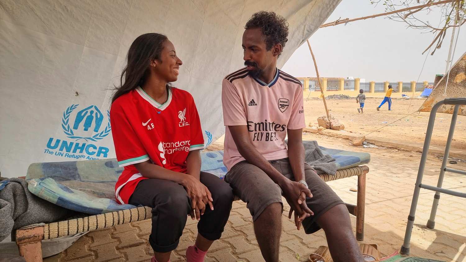 A young woman in a red football shirt smiles as she talks to a man sitting next to her under a UNHCR tarpaulin.