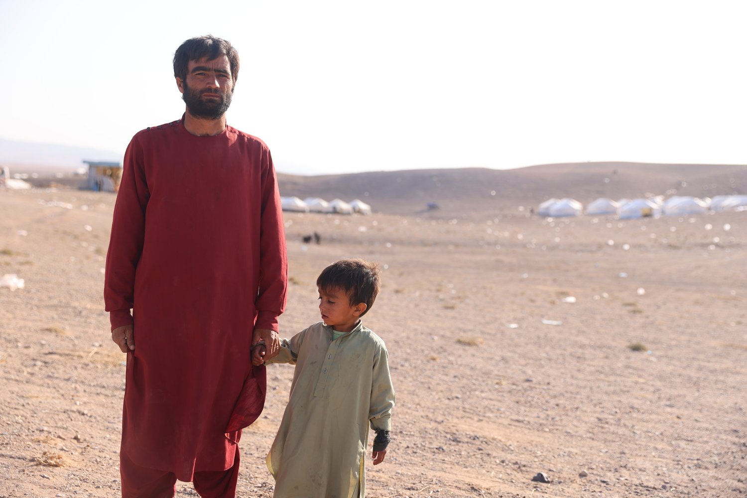 A father and young son stand in a barren landscape with rows of tents in the background.