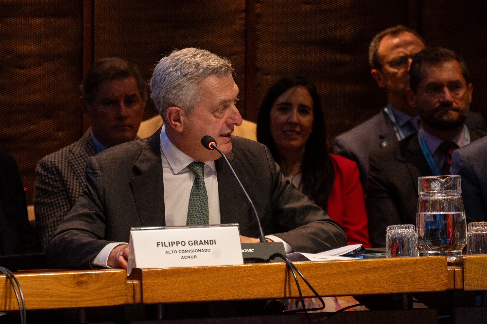 Filippo Grandi speaking at a desk with a microphone, a name placard in front of him and other people behind him.