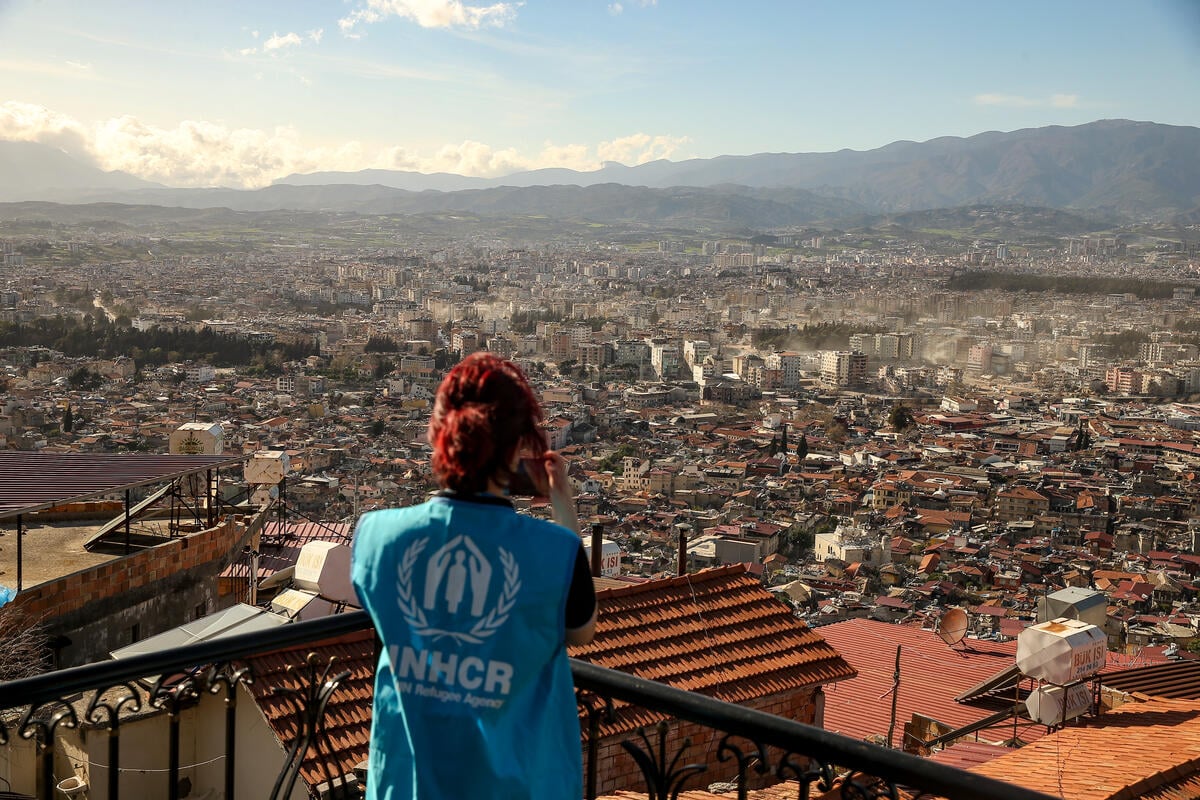 A woman in a UNHCR vest takes a photo of a city damaged by an earthquake.