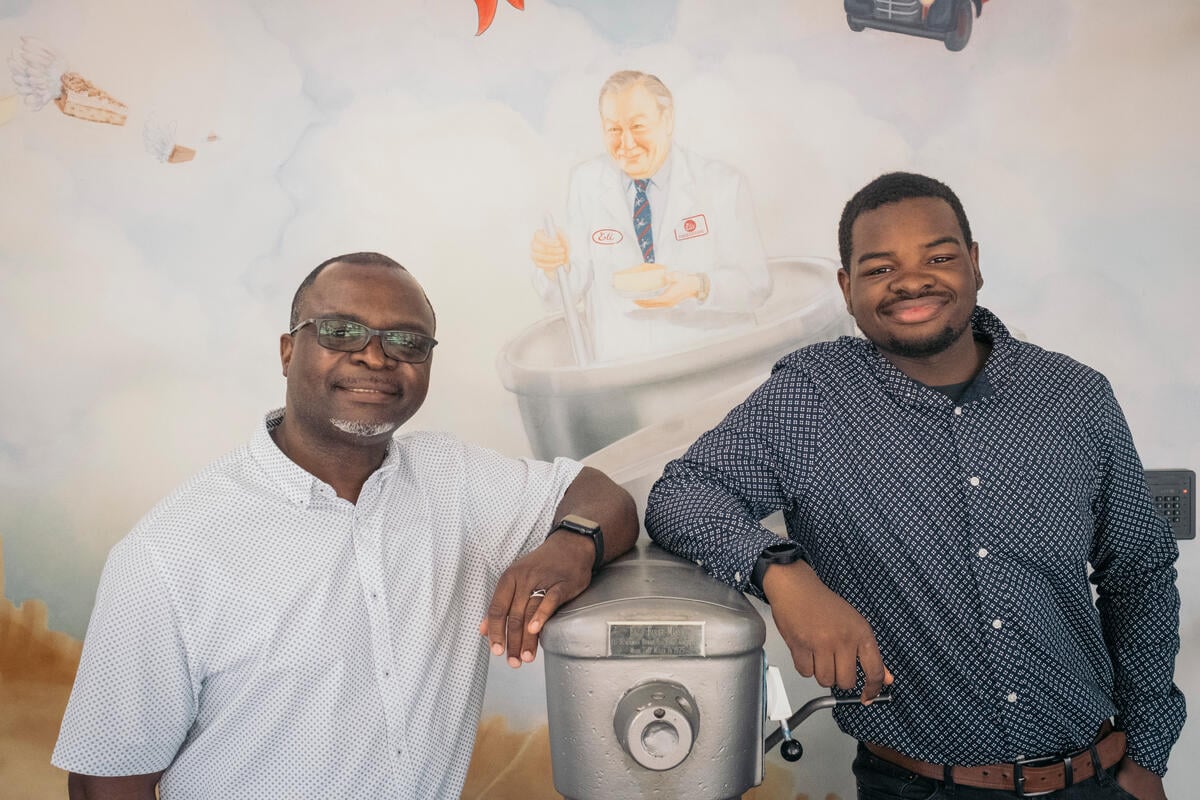Two men stand either side of an industrial cake mixing machine in front of a mural-painted wall