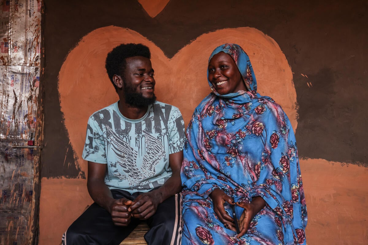 A woman and a man sit together, smiling at each other, in front of a heart-shaped wall painting