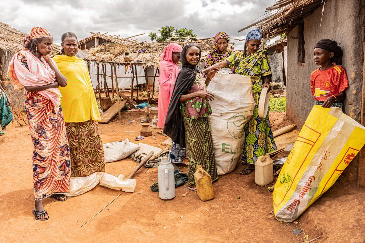 A group of women in colourful dresses stand outside a mud wall hut as they fill two large sacks with belongings